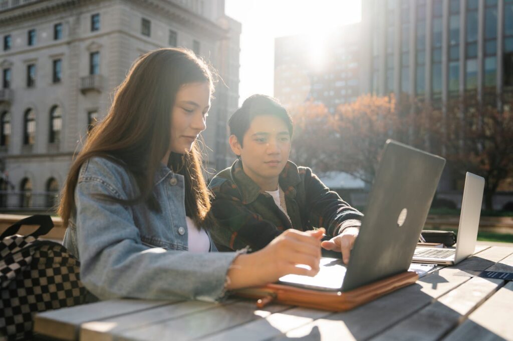 Two college students study together outdoors with laptops at a campus table.